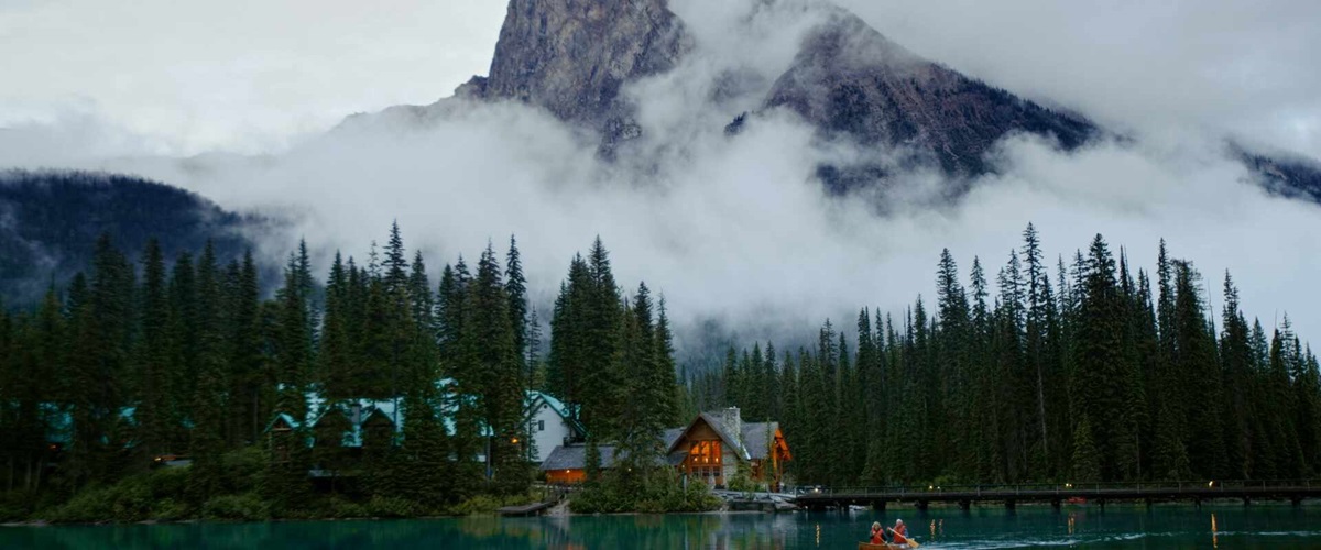 Yoho National Park - Emerald Lake. Wide shot of couple canoeing on calm lake in early evening, mountain and lights glowing in lodge in background